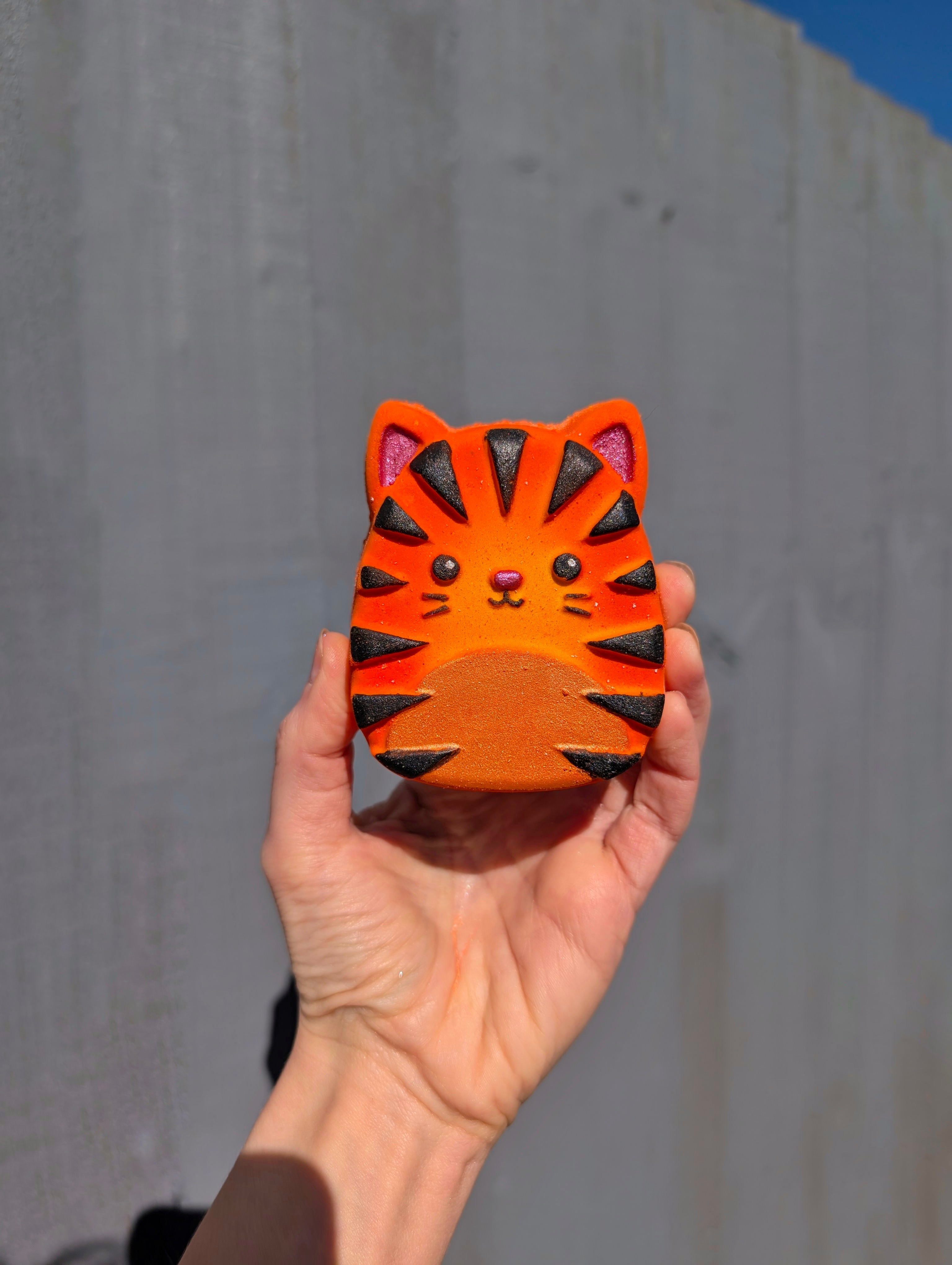 Hand holding a small orange Squishmallow tiger-shaped bath bombagainst a grey wall.