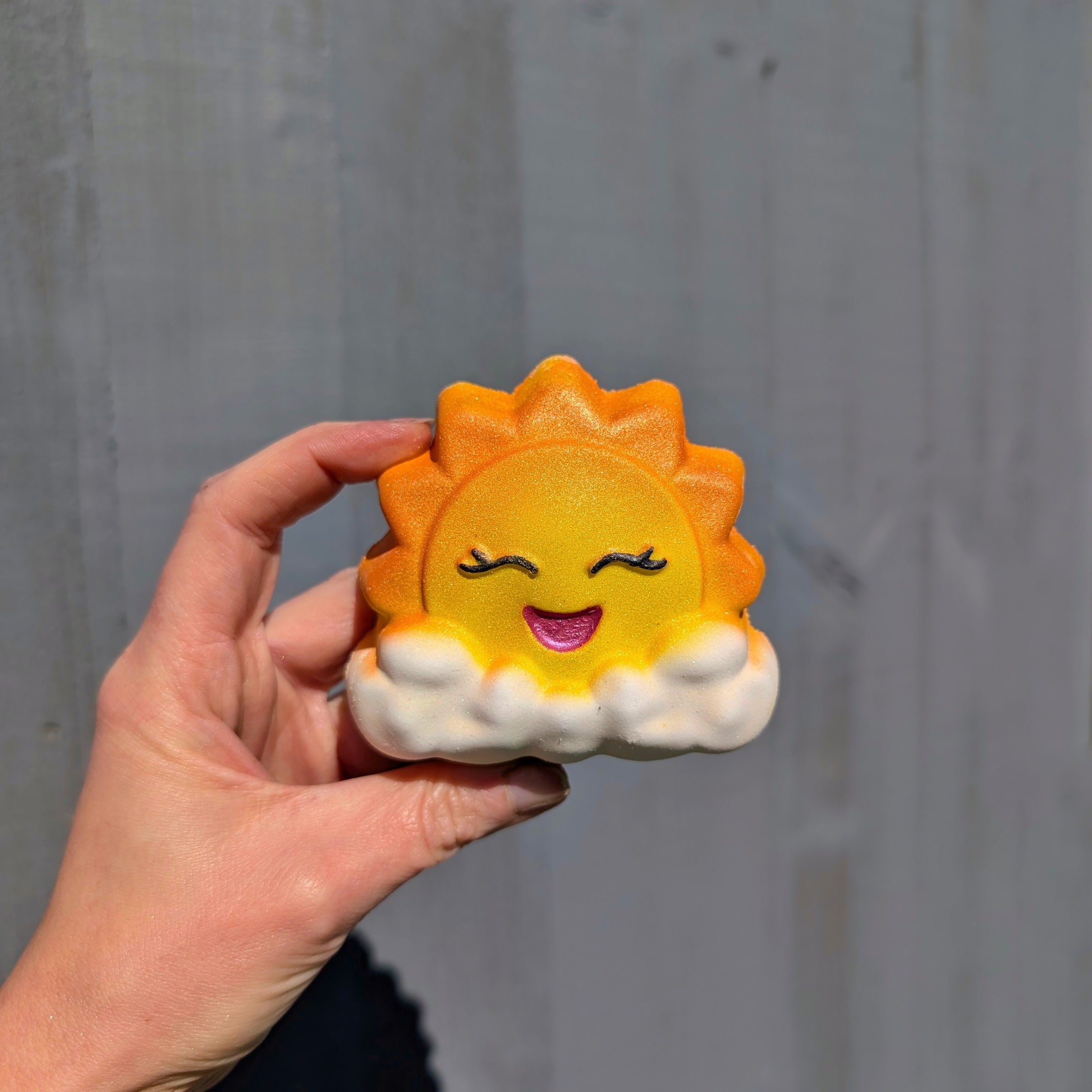 Hand holding a bath bomb shaped in a sun bursting out of the cloud, with a grey background.