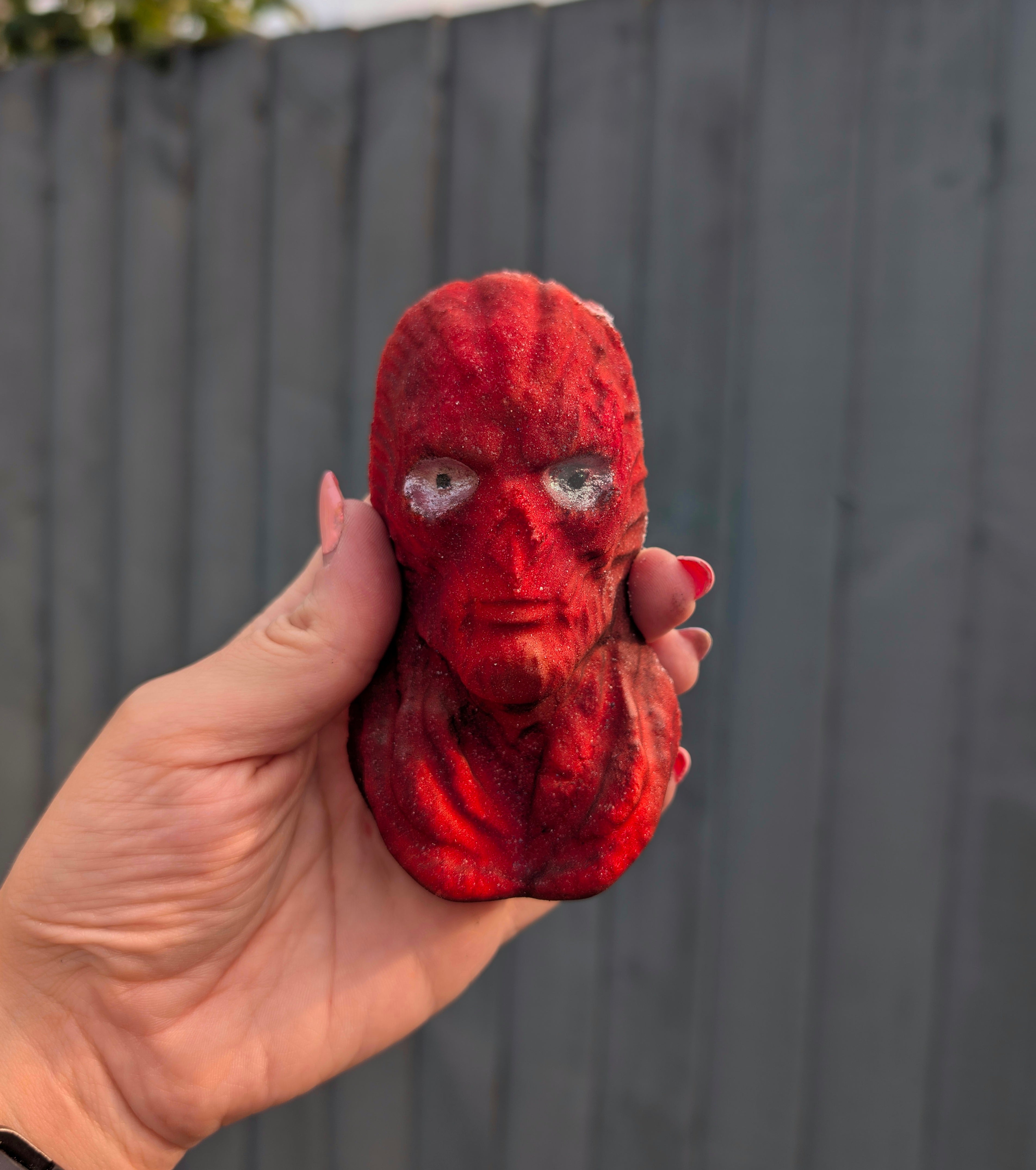 Vecna shaped bath bomb with a textured surface held by a hand against a grey wooden fence.