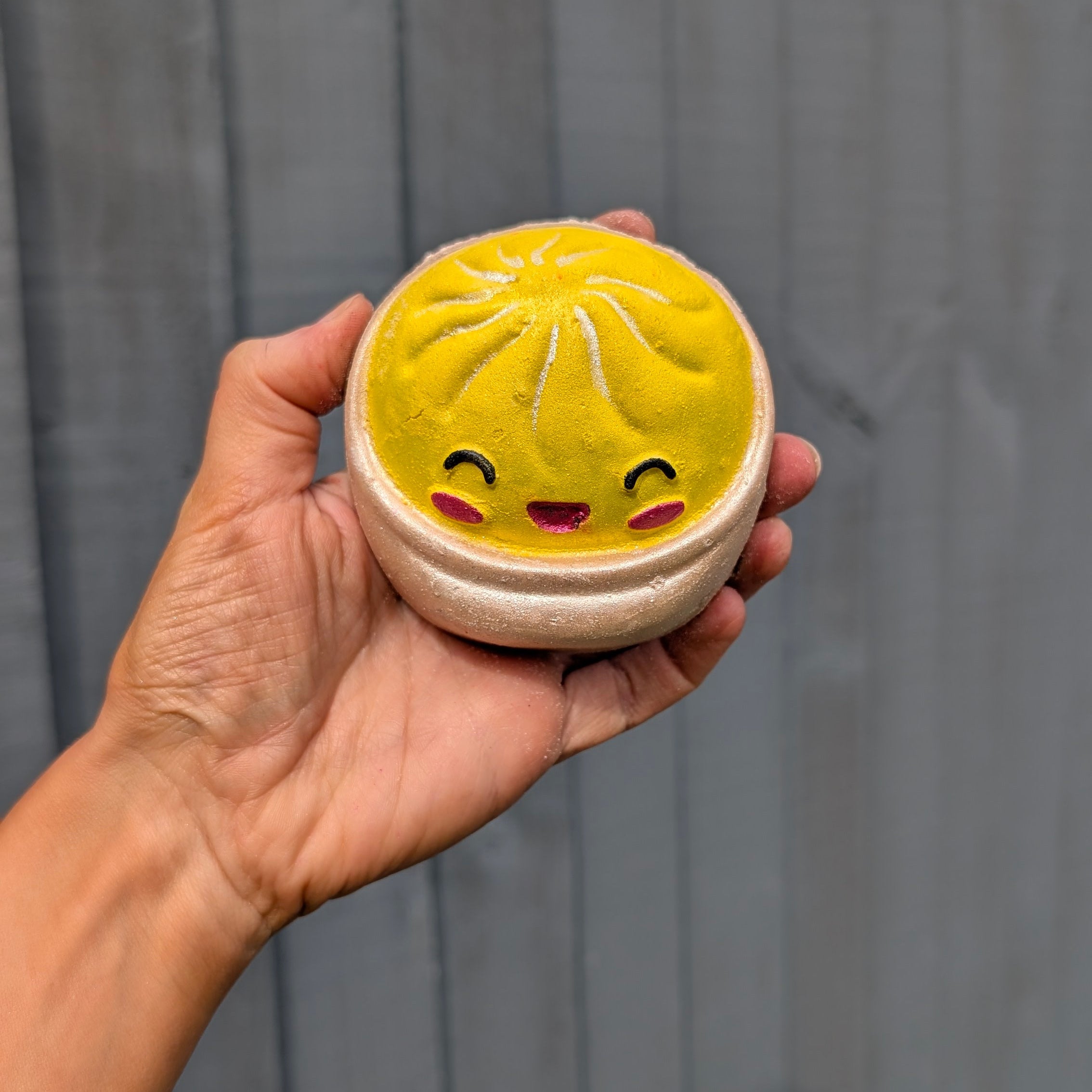 A hand holding a round dumpling bath bomb with a cute smiling face, set against a dark grey wooden plank background.