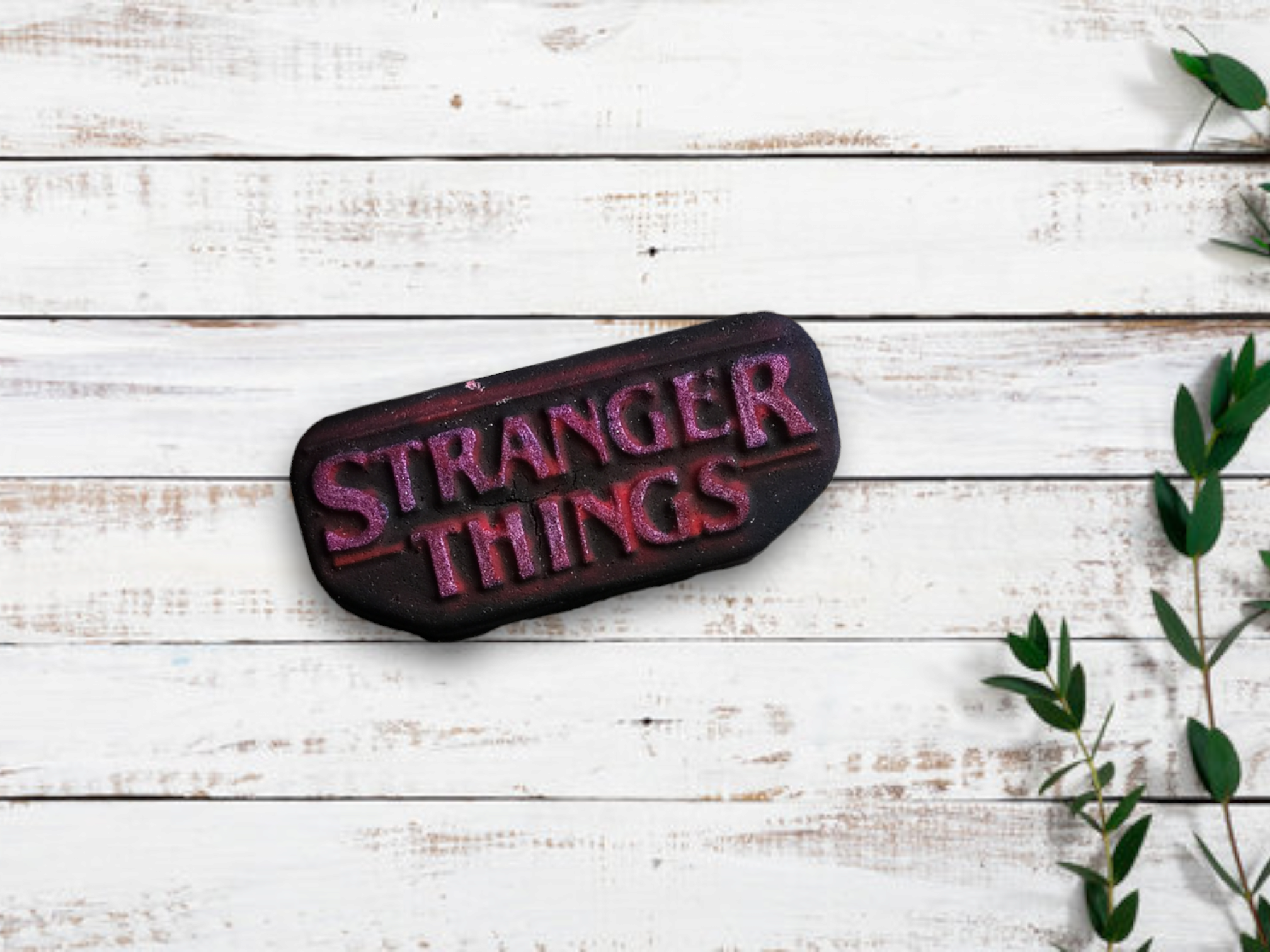 A bath bomb with 'STRANGER THINGS' text on it, placed on a wooden surface, with a few leaves of a plant to the side.